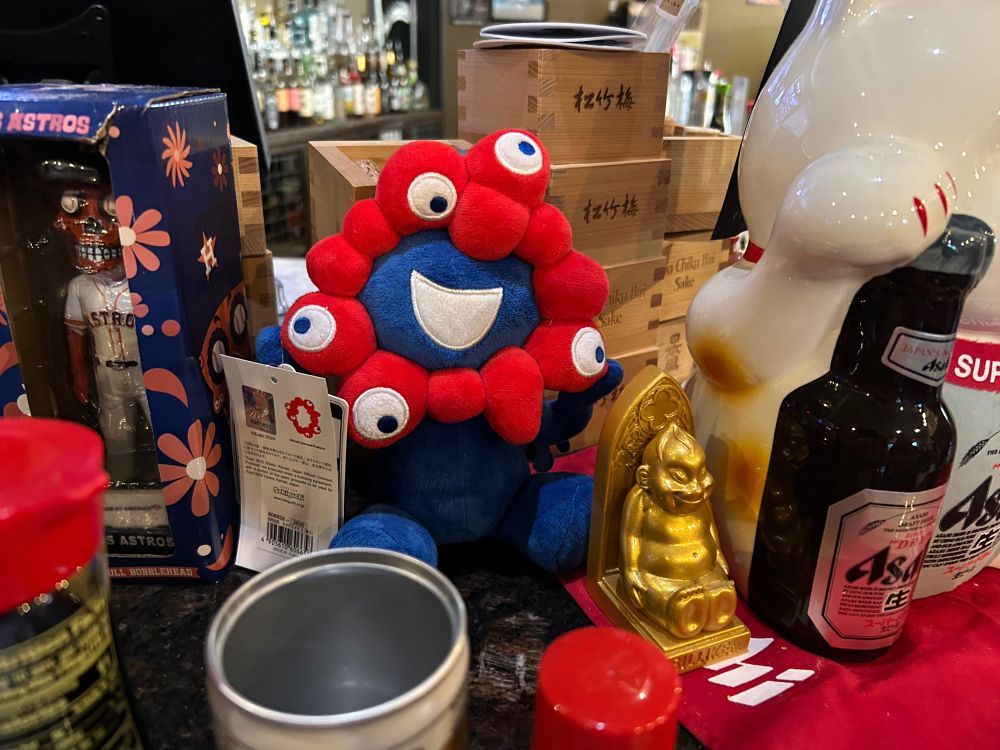 The Expo 2025 Osaka mascot next to an Astros sugar skull bobblehead, on a bar at a Japanese Izakaya in Houston.