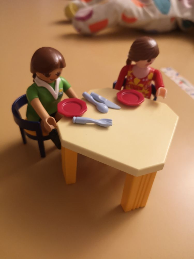 2 playmobil assises sur des sièges aux montants bleus, avec assise jaune et accoudoirs et installées à une table octogonale dans les tons jaunes sur laquelle sont dressés quelques couverts. 