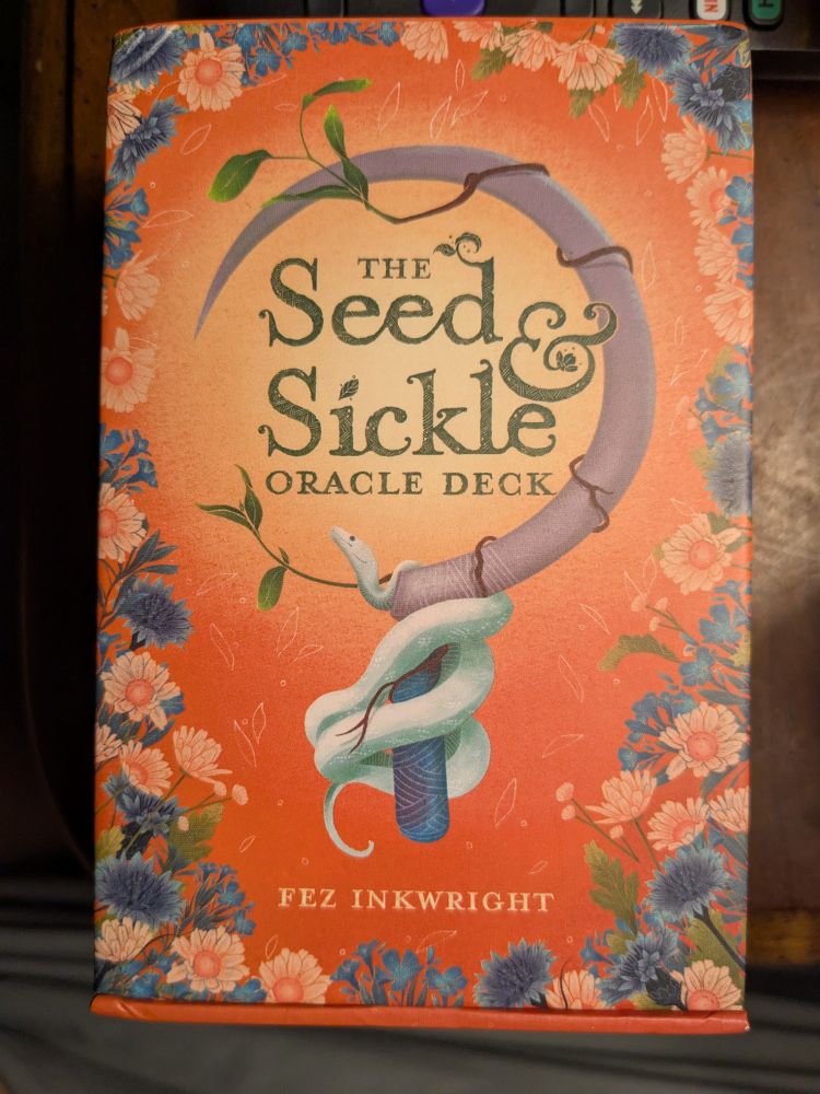 Image of an orange oracle deck box. Flowers frame an image of a white snake winding around a sickle handle while a vine circling the blade. 

The Seed & Sickle Oracle Deck by Fez Inkwright 
