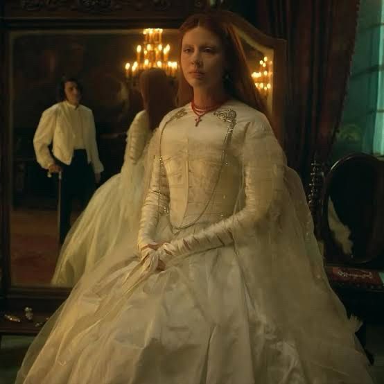 Photo of Mia Goth as Elizabeth in Frankenstein (2025) in her wedding gown costume; an out of focus Oscar Isaac as Victor Frankenstein can be seen reflected in the mirror behind her