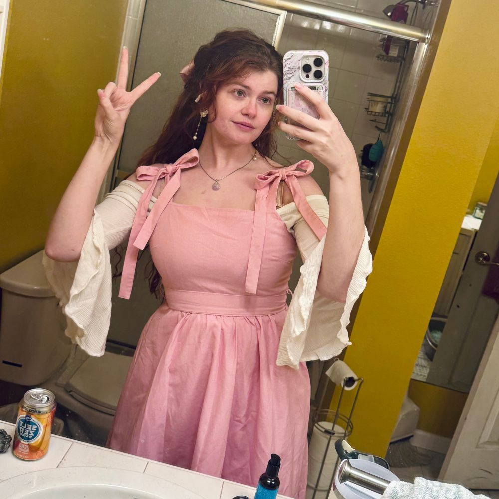 Image of Morgan, a woman with long red hair and a pink pinafore dress over a white off-shoulder blouse. She is taken a mirror selfie with one hand flashing a peace sign 