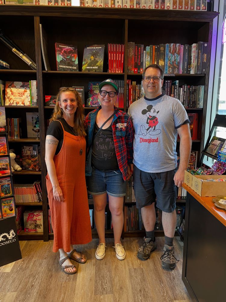 Jen, Andrea, and Blair standing together and smiling at the camera in front of a wall of TTRPG books. 