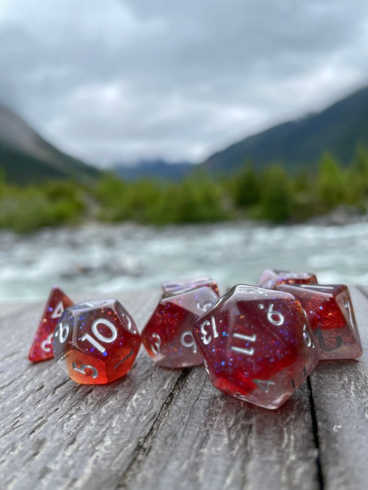 a set of sparkly red and clear polyhedral dice in a very clean focus, on a background of a wooden surface and a river with some green trees in blurry background