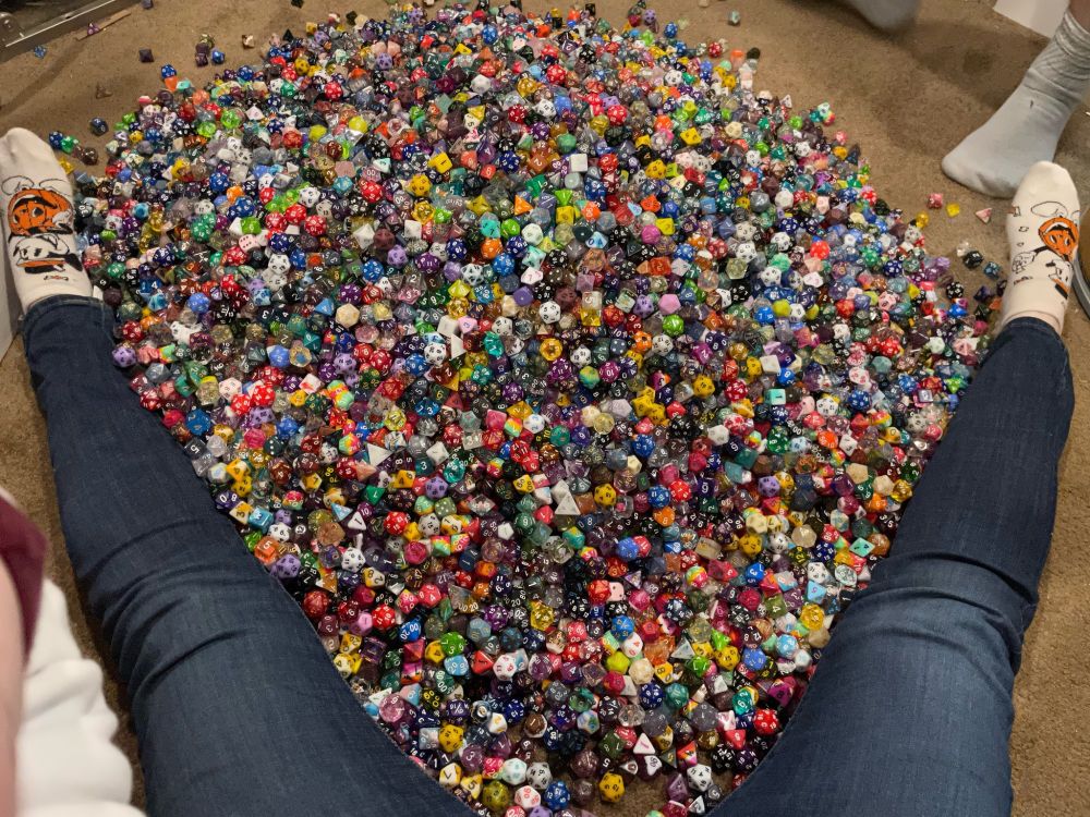 a giant pile of dice in between a person's legs; the photo is taken by the person sitting on the ground, to show just how many dice there are (it is many)