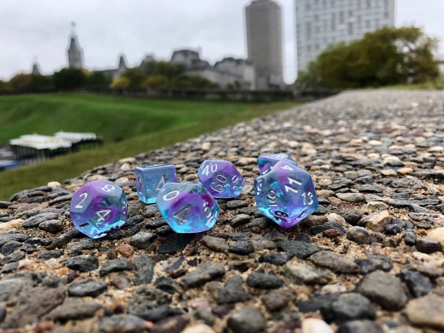 a set of polyhedral dice, in see through clear blue with purple swirls, on a cobbled wall with blurred buildings in the background