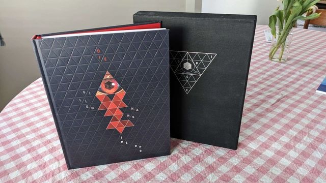 the deluxe edition of Die the RPG, with a textured faux leather black cover, and a red triangle with an eye inside it, over an open net made of many red triangles; next to it is a black slipcase with a silver geometric triangle reading DIE RPG
