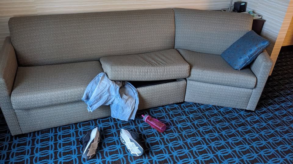 a very hard looking couch, with one cushion pulled up slightly and a pair of jorts dangling from beneath it; on the floor is a pair of shoes and a water bottle, to suggest that the couch is a mimic that has eaten a person