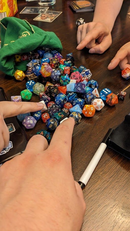 four hands pointing at a pile of assorted gaming dice on a wooden table