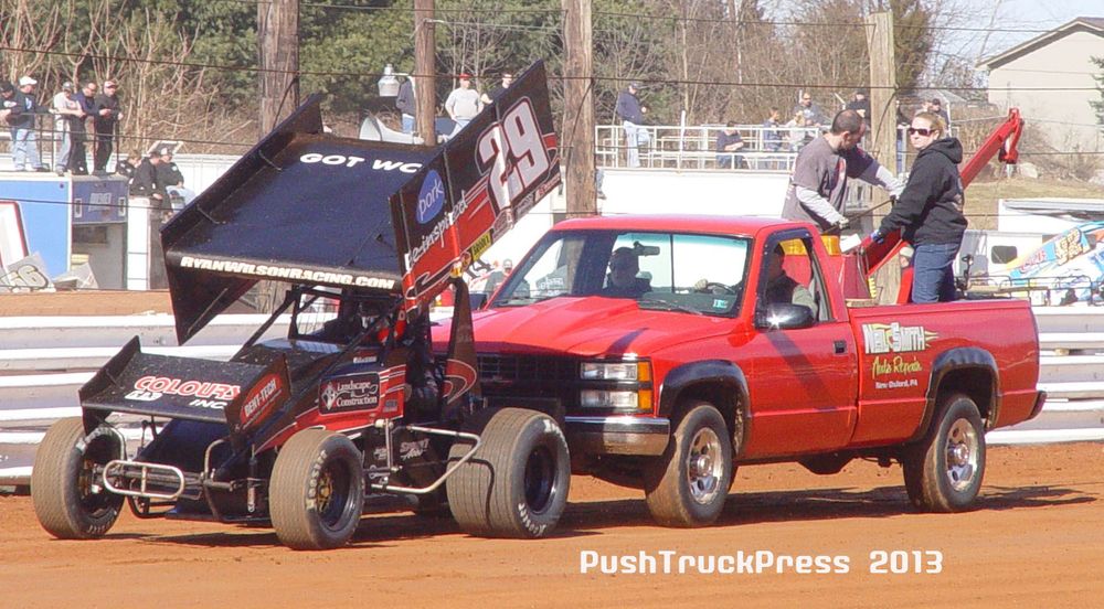 A red pickup truck push-starting a black winged sprint car on a dirt track