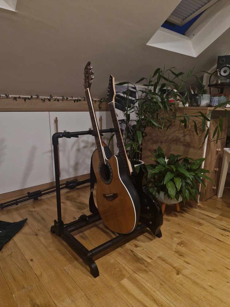 Image of a room with a slanted ceiling, the floor is wooden, white cupboards are visible in the background. There's plants and a desk in the background to the left. In the front is a black stand with two guitars propped on it, one timber acoustic with a black round back, the other electric with a slim curved solid body in a glossy black finish.