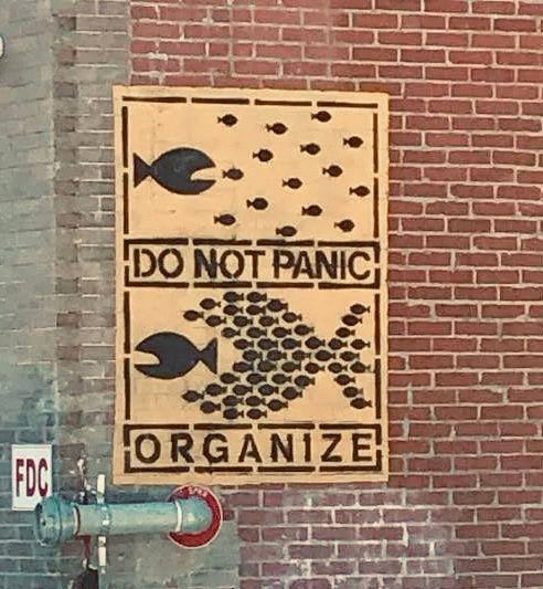 Painted sign on red brick wall--black stenciled art and text on yellow background: top of sign is large fish w open mouth going after a group of individual small fish--the text underneath says DO NOT PANIC: at the bottom of sign a much larger group of small fish in formation to make a very large fish with an open mouth pursuing a big fish and about to swallow it--the text underneath says ORGANIZE. 
