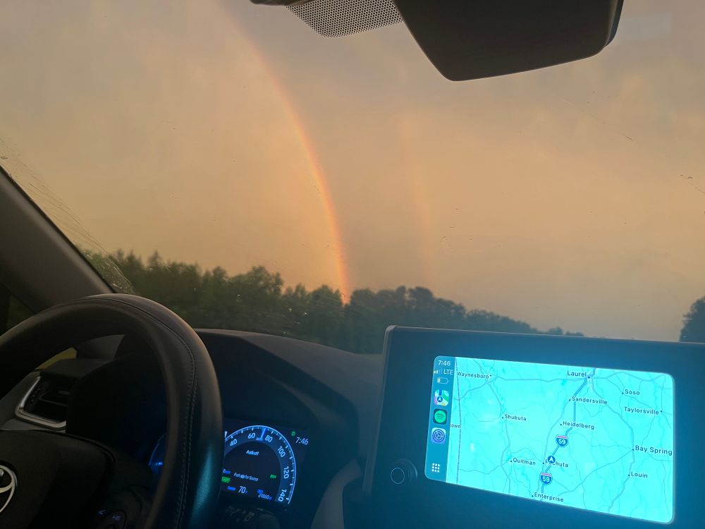 the view from the passenger seat of a car showing the steering wheel and dashboard with GPS and a double rainbow through the window. 