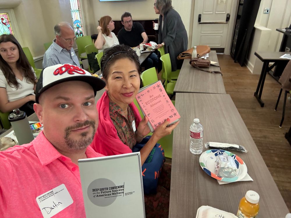 a white male wearing a baseball cap and holding a “deep south convening”  notebook sitting next to an asian female holding a pink “ecotone” booklet sitting at a long conference table with paper plates and bottled drinks and various people in the background. 