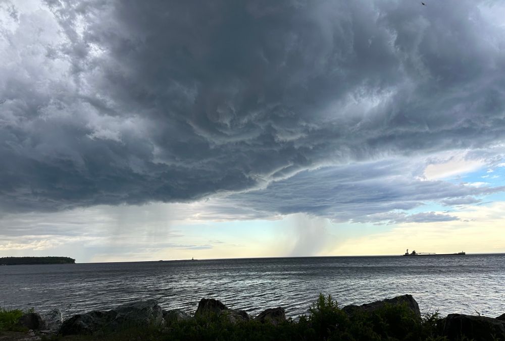 Thick get clouds on top of photo, waves of grey rain fall on silver Lake Superior water. A freighter is on the right horizon, the iron ore dock is on the left horizon 
