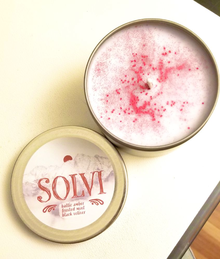 a candle with the lid off, showing red glitter on white wax like blood on snow. the label reads ‘SOLVI: baltic amber, frosted mint, black vetiver’