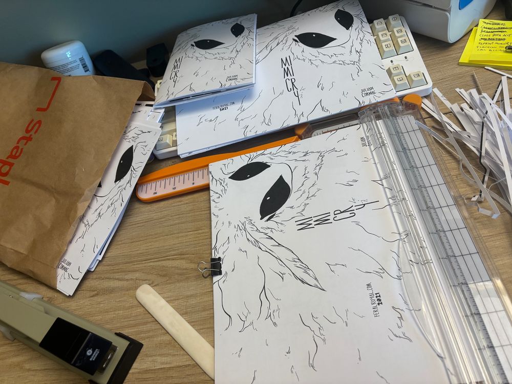 A photo of a desk being used for comic zine assembly. Some are stapled and waiting to be folded, one is in the process of being trimmed.