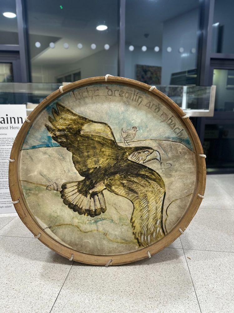 Hand-painted traditional drum featuring an artistic depiction of a large eagle in flight, with detailed feathers and a small crowned wren perched on its back. The drum is encircled by a handwritten Irish inscription reading 'Inbhe Dreolin Ard Mhacha,' translating to 'The Wren of Armagh,' referencing Irish folklore and cultural symbolism. The artwork emphasises the contrast between the powerful eagle and the small but significant wren, reflecting themes of strength, wisdom, and legacy. The drum is displayed in a modern museum or cultural exhibition setting, with a backdrop of glass and informational plaques, highlighting its cultural and historical significance.