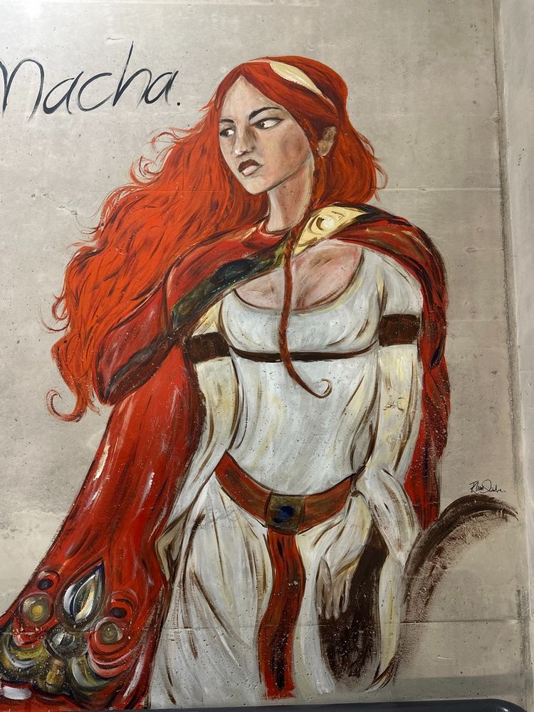 Painting of Macha, a legendary figure from Irish mythology, depicted as a powerful and elegant woman with flowing red hair. She wears a white dress cinched with a belt, complemented by a richly decorated red cloak adorned with intricate patterns resembling peacock feathers. Her expression is resolute, with a piercing gaze that reflects her strength and sovereignty. Macha is one of the three aspects of the Morrígan and is associated with war, fertility, and the land. The artwork captures her commanding presence and mythical significance, displayed against a minimalist background to emphasize her figure.