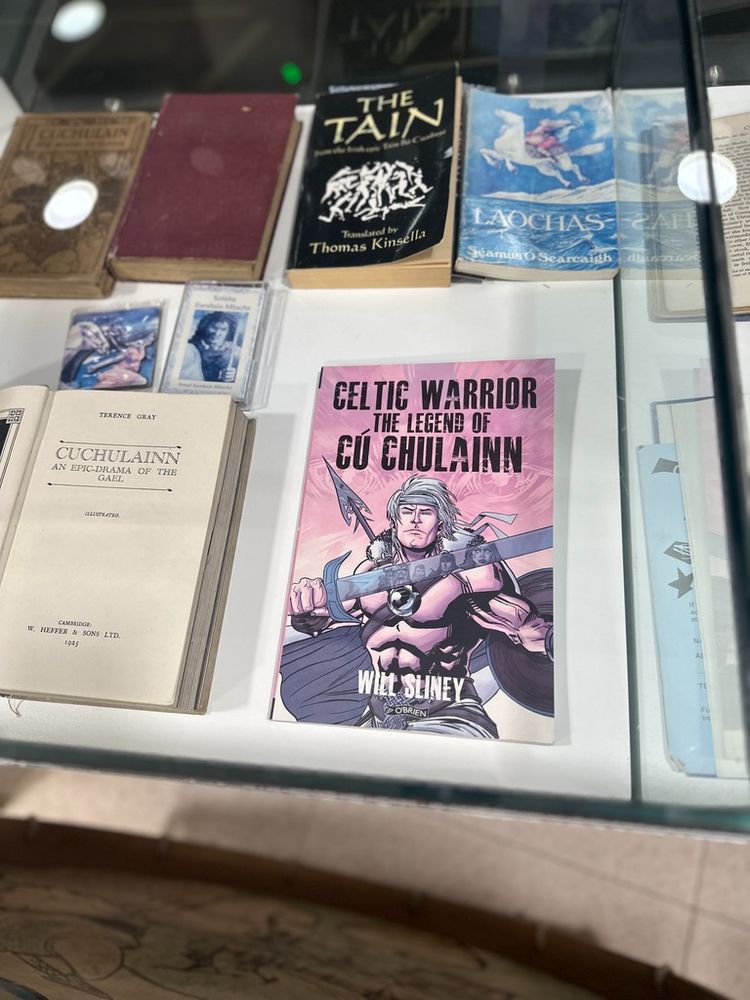 Display case featuring a collection of books and literature related to Irish mythology and folklore, with a prominent comic-style book cover titled 'Celtic Warrior: The Legend of Cú Chulainn' by Will Sliney, depicting a muscular warrior holding a sword and shield in dynamic artwork. Surrounding items include classic texts like 'The Táin' translated by Thomas Kinsella, 'Laochas' by Séamus Ó Searcaigh, and vintage editions of works exploring the mythology of Cú Chulainn, such as 'Cuchulainn: An Epic-Drama of the Gael' from 1925. The arrangement emphasises the blend of traditional and modern interpretations of Irish legendary tales, showcasing the enduring cultural significance of heroic figures like Cú Chulainn.