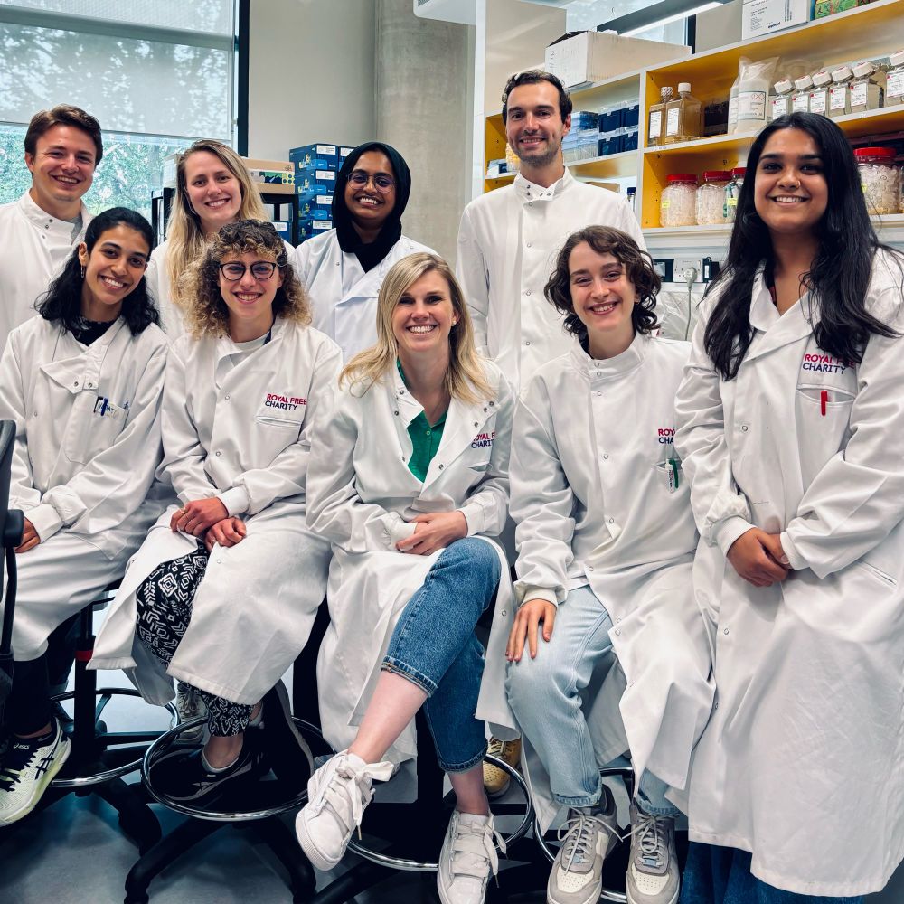 A group of academics wearing white lab coats in a research Institute. Some are sitting. Some are standing. Everyone is smiling. 