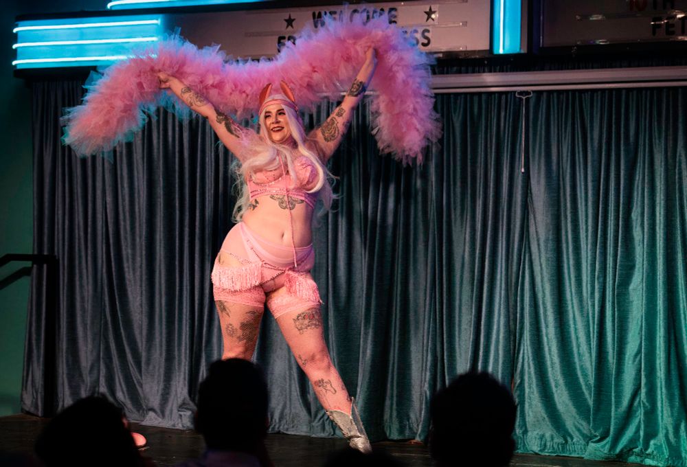 Ryder Strong wears a blonde wig and pink leather pony play ears with long pint reigns. She is wearing a pink rhinestoned bra with matching underwear and a pink fringe shimmy belt. She is holding a large pink tool boa above her head in front of a blue curtain.