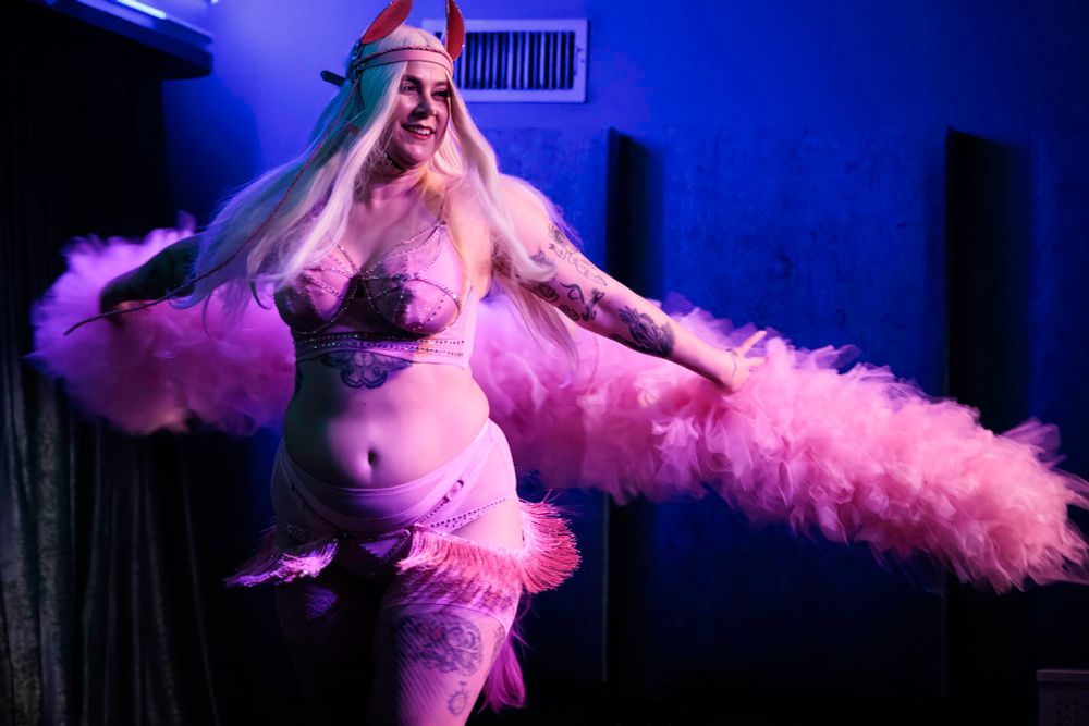 Ryder Strong wears a blonde wig and pink leather pony play ears with long pint reigns. She is wearing a pink rhinestoned bra with matching underwear and a pink fringe shimmy belt. She is spinning with a large pink tool boa behind her while she smiles at the crowd.