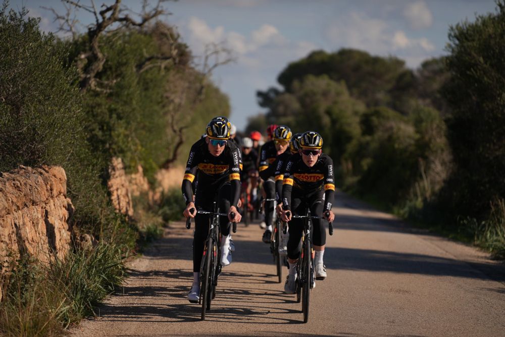 Group of Ineos and Lotto-Kernhaus riders front on riding down a road two abreast in the countryside of Spain.