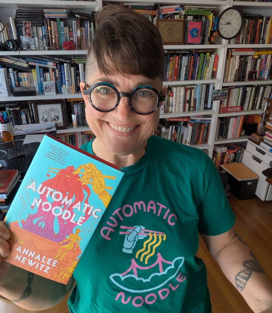 I am smiling, standing in front of my bookshelf, wearing a green t-shirt that says "Automatic Noodle" and holding up a copy of my book. 