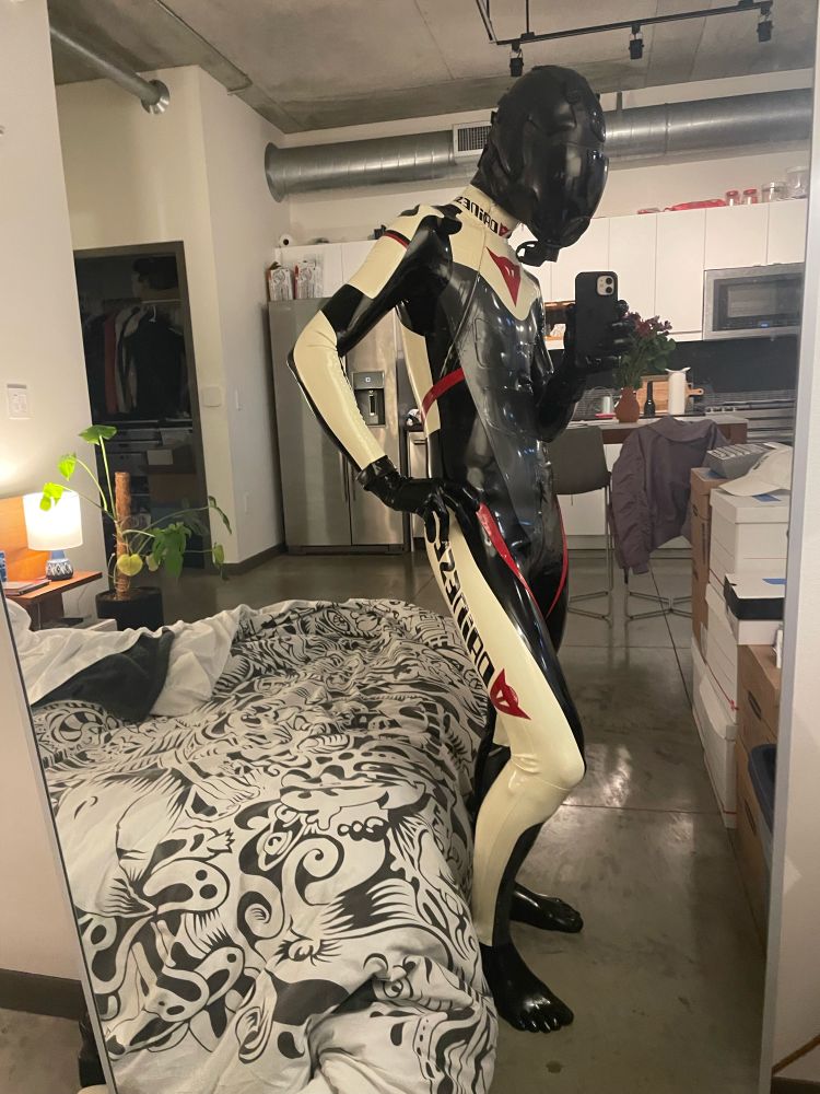 A drone in a reflective mirror is wearing a black and white racing suit with a studiogum hood and msa gasmask.

Side profile with right hand on its hip. 