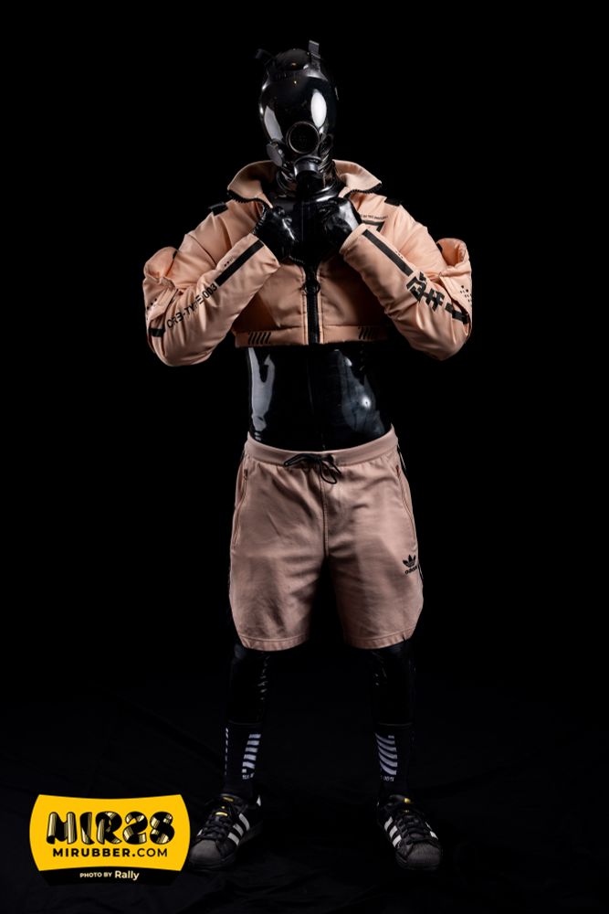 Rubber drone, is that tech or scaly footwear? Wearing a black latex catsuit under pink tech wear with an MSA gasmask, Adidas Superstars, and socks with white strips. Posing holding the collar of its crop top jacket