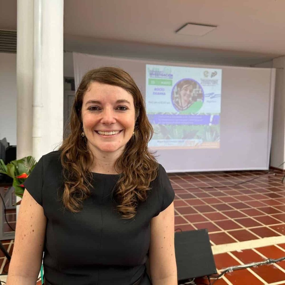 Dr. Rocío Deanna with shoulder-length wavy brown hair wearing a black sleeveless top stands in front of a presentation screen. The screen displays a colorful slide with circular diagrams and text. 