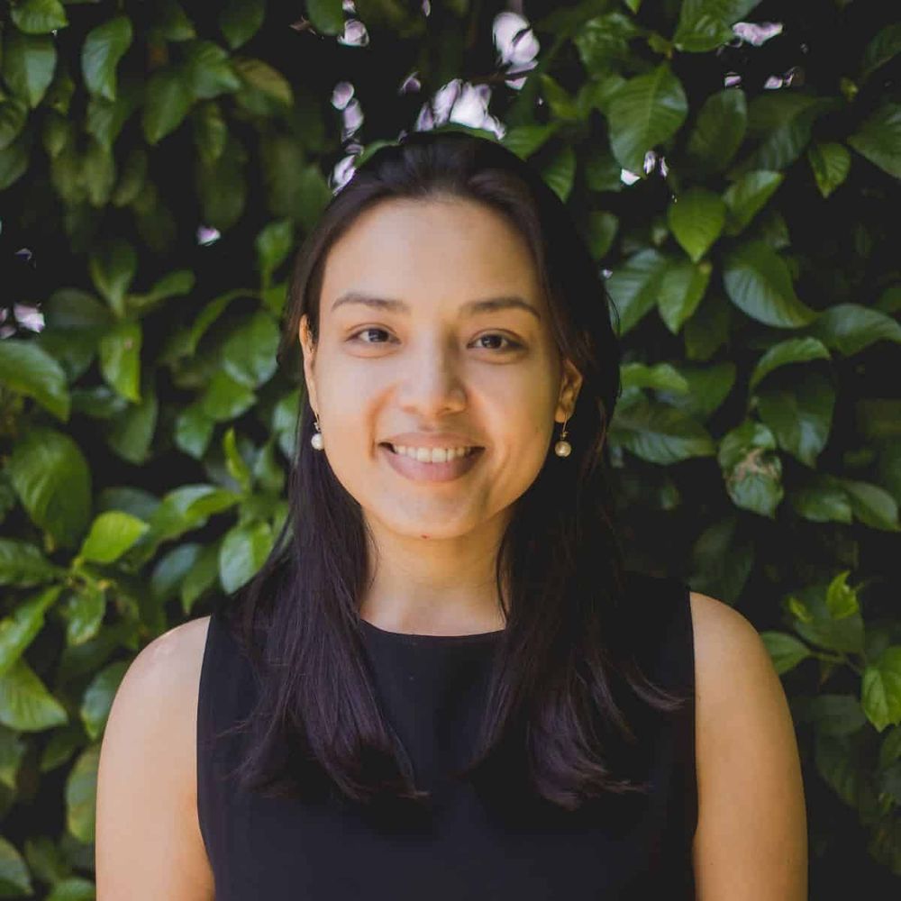 Dr. Juliane Ishida, a smiling woman with long dark hair wearing a black sleeveless top and pearl earrings, photographed against a lush green backdrop of leafy plants with small pink flowers.