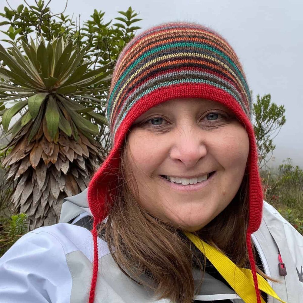 Dr. Laura Lagomarsino smiling at the camera while conducting fieldwork in a tropical environment. She wears a colorful striped knit hat with red trim and a light-colored field jacket with yellow straps. Behind her is a distinctive tropical plant with large, paddle-shaped leaves arranged in a rosette pattern, characteristic of high-altitude Andean vegetation.