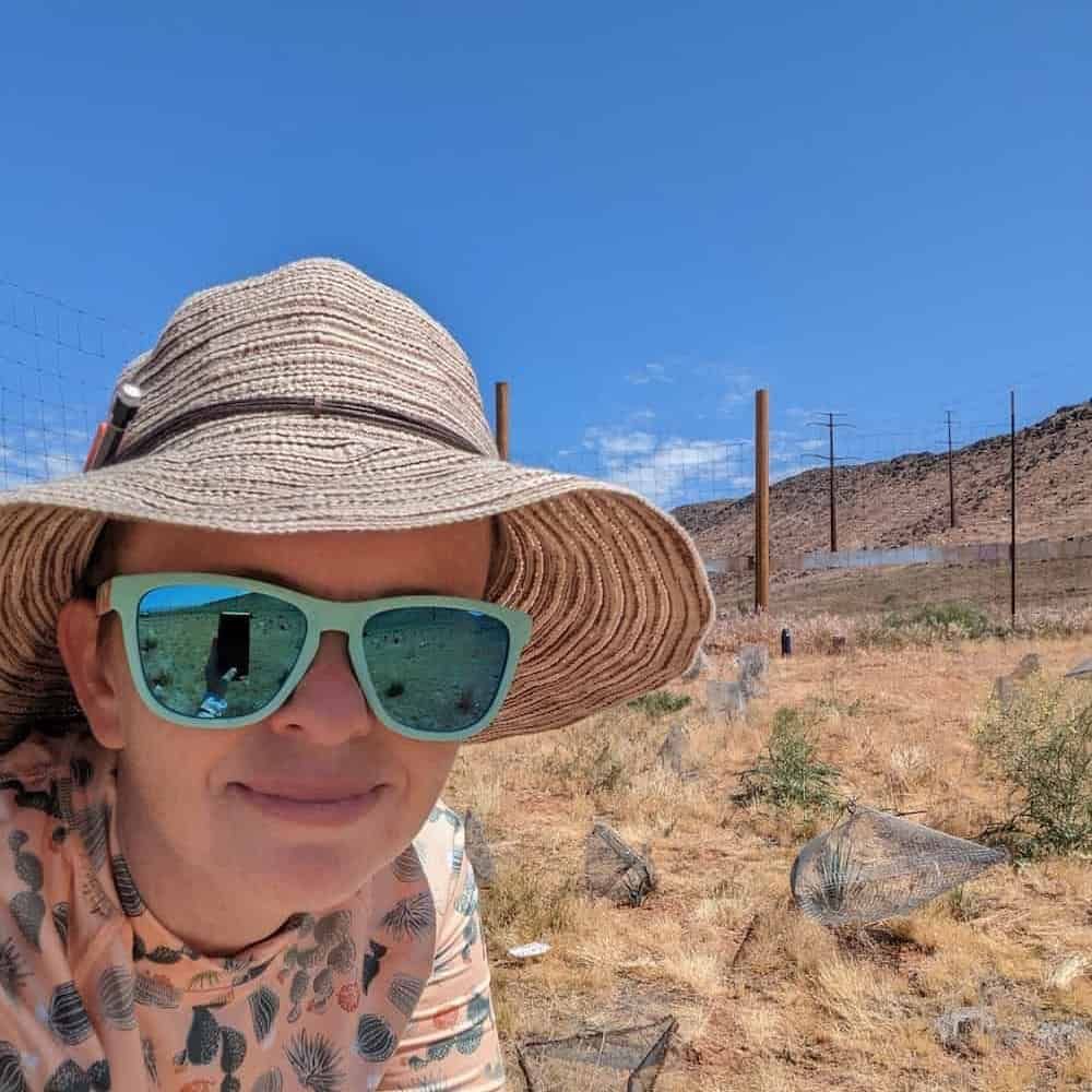 Karolina Heyduk, taking a selfie on fieldwork. She is in a dry landscape with brown, parched vegetation. Above is a strikingly blue sky. She is looking into the camera, wearing a cactus-themed top, a light brown sun hat and sunglasses. In one lens of the sunglasses is a reflection of her hand taking the selfie. I've remembered I'm colour-blind, so take any references to brown with a pinch of salt.