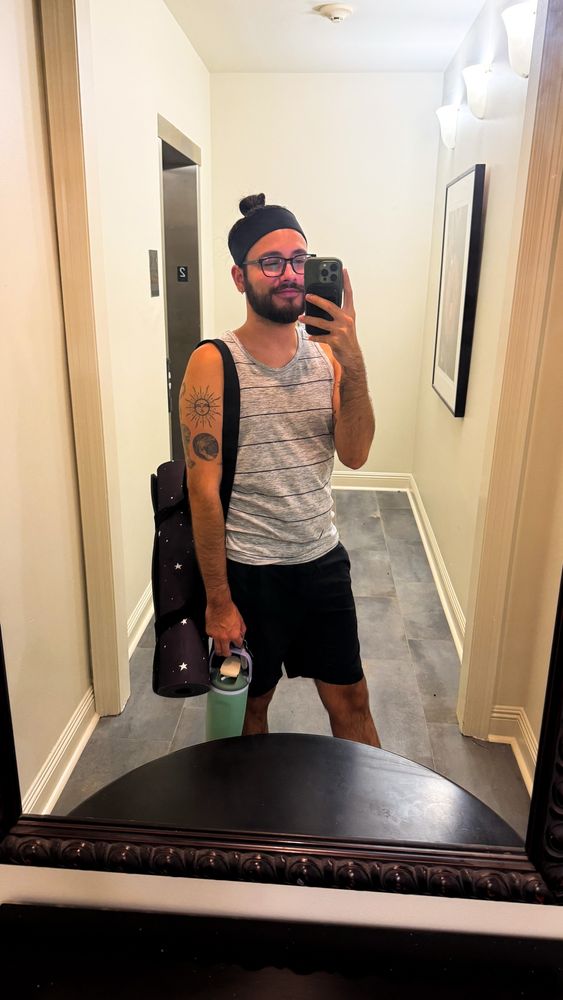 Mirror picture of myself before yoga class in the elevator hallway. My hair is in a man bun and I have an athletic headband on. Wearing a grey tank top with black stripes and black athletic shorts. I have a black yoga mat with white starts hanging over my shoulder and I’m holding my water bottle. 