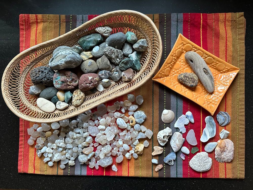 Variety of ocean rocks in an oval basket with textured surfaces and a range of colors including red, green, black & tan. Large oval pile of small mostly white agates. Ocean-worn shell fragments & sea glass. Rectangular orange ceramic dish with a small piece of smooth driftwood next to a speckled orange and brown rock. All objects arranged on top of a striped, multi-colored piece of fabric in autumn hues. 