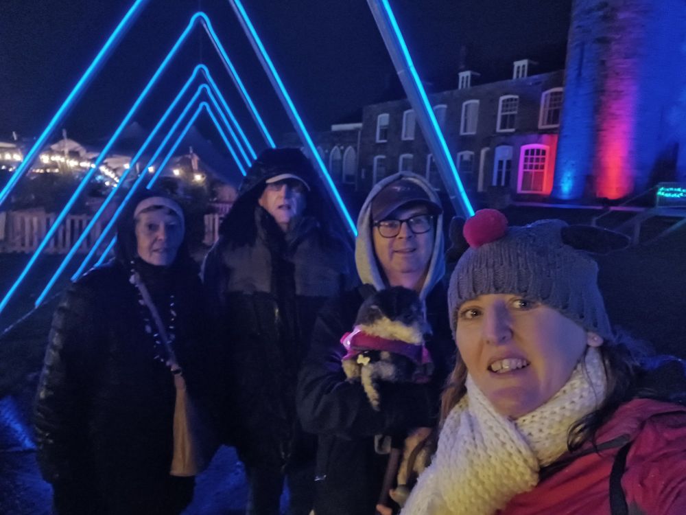 Two couples are wrapped in winter clothing, standing beneath an arch made of blue lights.