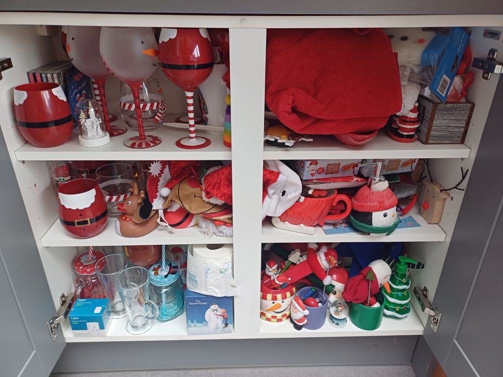 An open cupboard with three shelves full of Christmas themed objects such as Santa and snowman glasses, a red gravy boat and a Christmas tree soap dispenser. 