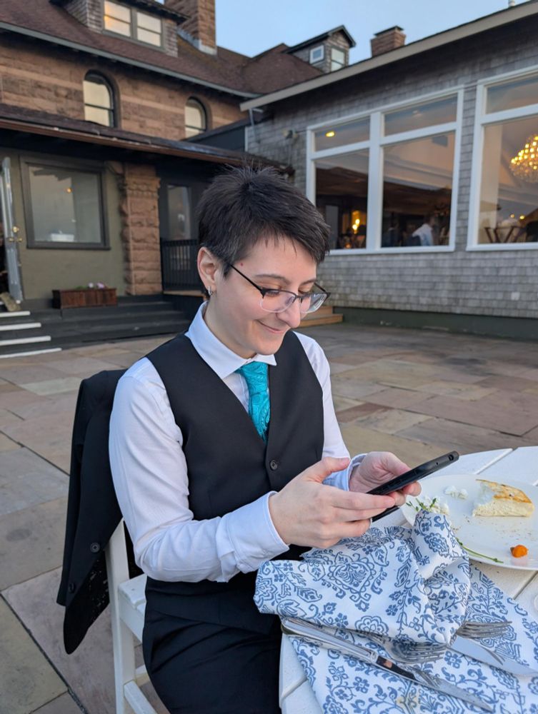 I'm sitting at a table outside, distracted on my phone and smiling. Wearing a black suit, white button down shirt and teal brocade tie.