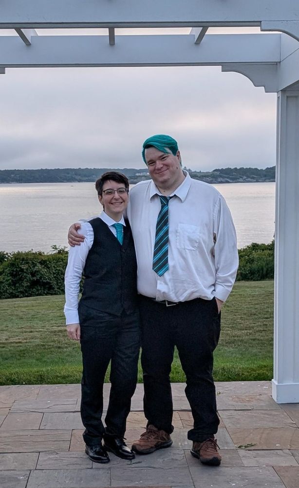 Me and my spouse standing under a white wooden arch in front of the ocean. We're both smiling. It is wearing a white button down shirt, teal striped tie, and black pants. I am wearing a black suit, white button down shirt, and teal brocade tie.