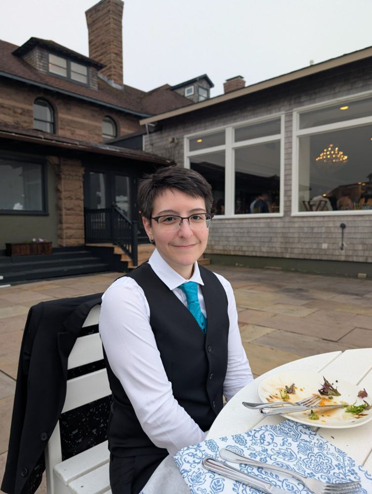 Me sitting at a table outside, wearing a back vest and pants, white button down shirt and teal brocade tie. I'm smiling at the camera.