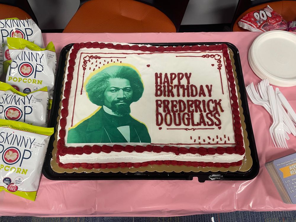 White sheet cake with red piping that reads “Happy Birthday Frederick Douglass” with an image of Douglass. 