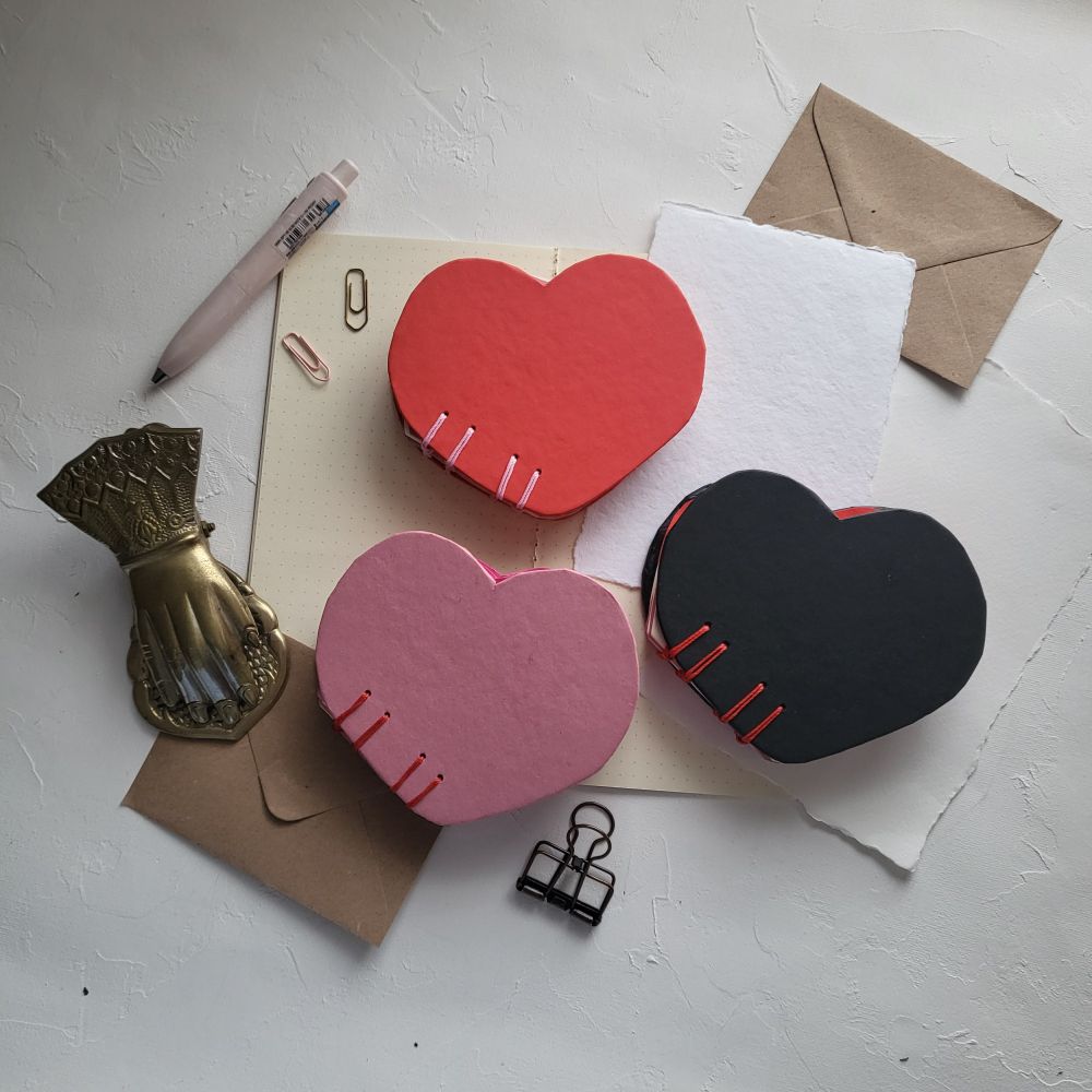 In the centre of the photo are three heart-shaped notebooks in the formation of a triangle. The top is a red heart-shaped notebook with pink coptic stitching with the bottom left of the formation, which is a pink heart-shaped notebook with red coptic stitching, and next to it is a black heart-shaped notebook with red coptic stitching. These notebooks are lying on top of kraft paper, white paper, and a6 sized envelopes. There are two paperclips, a pink pen and two book clips to help frame the notebooks. 