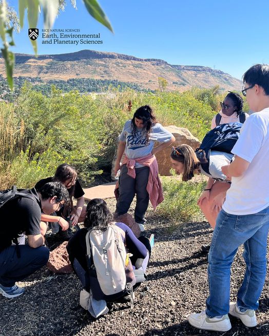 Students stand and squat around an interesting rock with mountains behind them.