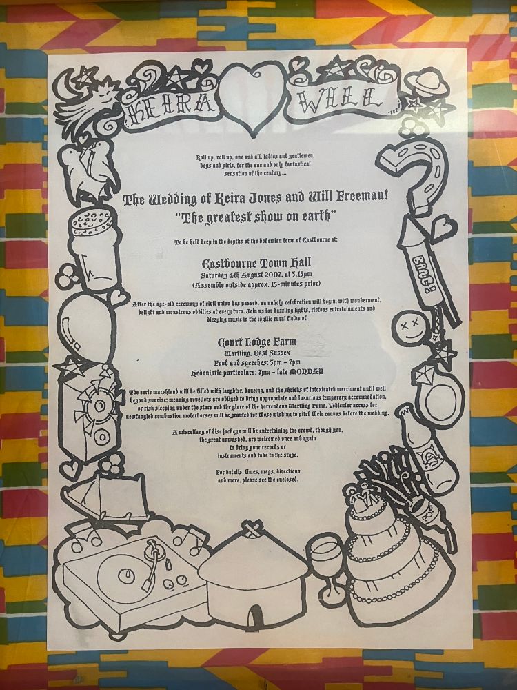 A photograph of a large back and white wedding invite, mounted on a wall and surrounded by multicoloured fabric.