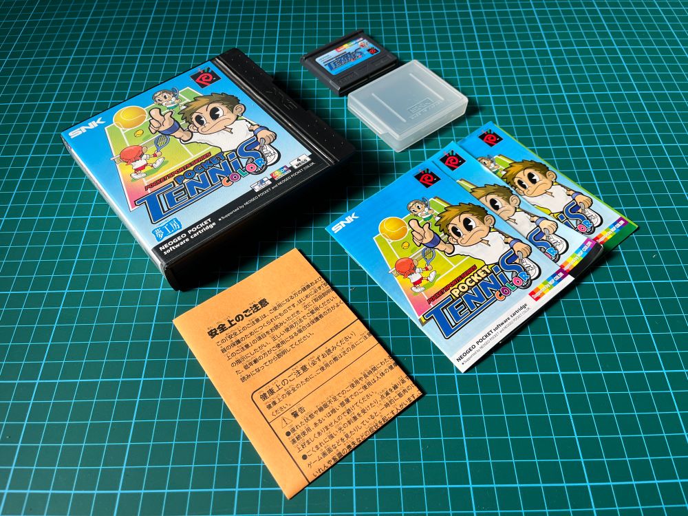 A boxed copy of the NeoGeo Pocket Color game Pocket Tennis Color, placed on a green cutting mat, with the manuals, orange registration card, cartridge and cartridge case displayed alongside the box.