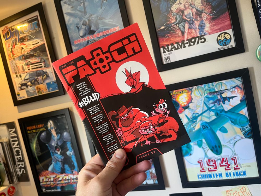 A hand holding up a copy of indie games magazine Patch, in front of a wall covered in framed arcade flyers.