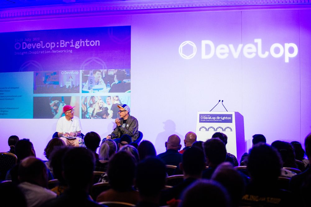 A photo from a few rows back in the audience at the Develop Brighton conference in 2013, with Will Freeman (left, pink cap) and Chris Charla (right, multicoloured cap) in discussion on stage. In the backdrop is a large logo for the conference, an empty lecture, and a projected screen showing a slide.