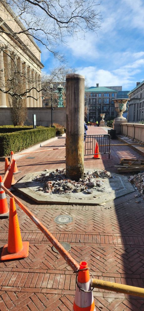 The western flagpole, with about 7-9 feet of wood left above ground. The base has been cleared away, and there's brick rubble around it. Orange cones block it off.