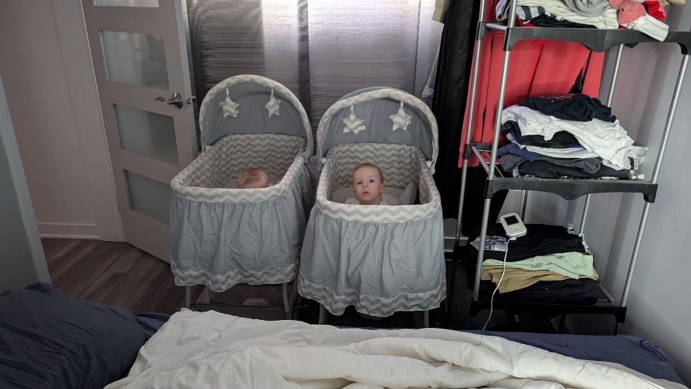 Twins in their bassinets.
One is looking straight at the camera while crawling, the other is also on her belly, but we just see the top of her head.